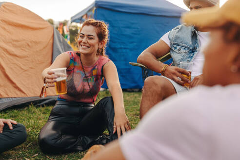 Joyful young woman raises her beer glass in a festive toast during a lively celebration at the summer festival. Young woman sitting at a campsite, surrounded by friends and festival goers. This photo has intentional use of 35mm film grain. - JLPPF02199