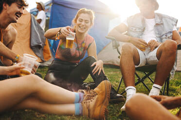 Happy young woman joyfully raises her beer cup in the vibrant atmosphere of a summer festival camp, joining her friends in celebrating and cherishing the moment. This photo has intentional use of 35mm film grain. - JLPPF02200