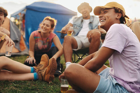 Group of young people celebrate and bond together at a summer festival camp, laughing, chatting and having fun over cold beers. This photo has intentional use of 35mm film grain. - JLPPF02202