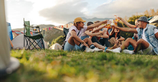 Group of young adults gather together, smiling and toasting with beer in celebration of a summer festival. Friendship, enjoyment and refreshment fill the carefree day as they bond over drinks. This photo has intentional use of 35mm film grain. - JLPPF02212