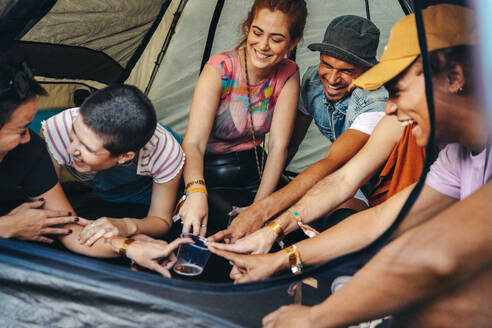 Friends gather in a festival tent, engaging in a fun and lively drinking game, adding an extra layer of excitement to their festival adventure. Group of young people having a good time at a summer festival. This photo has intentional use of 35mm film grain. - JLPPF02223