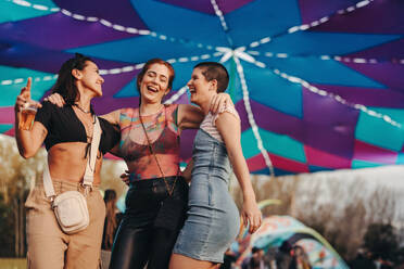 Group of friends joyfully immerses themselves in the festive atmosphere of a summer festival, sharing laughter, music, and unforgettable moments of fun. Three young women enjoying a festival party. This photo has intentional use of 35mm film grain. - JLPPF02233