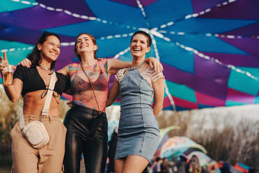 Three female friends laugh with happiness and excitement as they fully embrace the party vibes and vibrant energy of a music festival. Group of young women living their best lives at a summer festival. This photo has intentional use of 35mm film grain. - JLPPF02234
