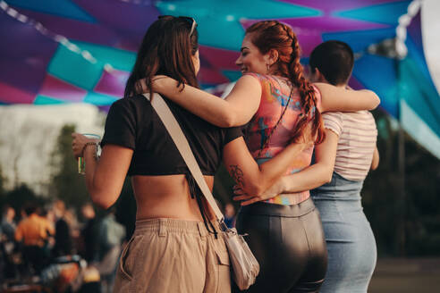 Three female friends come together in a warm embrace, their spirits lifted by the joyous atmosphere and immersive experiences of a blissful summer festival. This photo has intentional use of 35mm film grain. - JLPPF02236