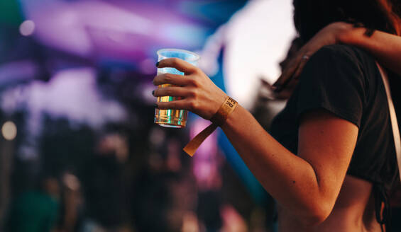 Woman holds her cup of beer, standing amidst the lively venue of a music festival, ready to immerse herself in the vibrant festivities. This photo has intentional use of 35mm film grain. - JLPPF02237