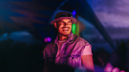 Happy young dj stands in a vibrant festival atmosphere, surrounded by neon lights and lively music. Man smiling and celebrating summer nightlife with excitement. This photo has intentional use of 35mm film grain. - JLPPF02239