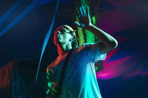 Young dj stands under vibrant neon lights on a festival stage, dancing and enjoying the music during a night concert. Young man creating a vibrant ambiance for the crowd at a summer music festival. This photo has intentional use of 35mm film grain. - JLPPF02243