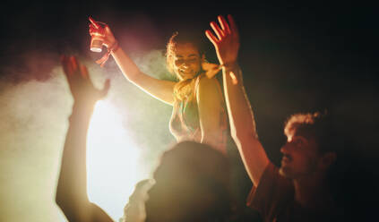 Young people joyfully celebrate and dance together under the vibrant lights of a summer music festival. Crowd of young people raising their hands and having the time of their lives in a festival atmosphere. This photo has intentional use of 35mm film grain. - JLPPF02255