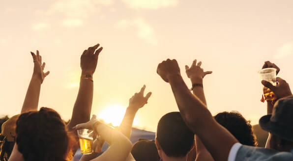 Vibrant crowd raises their hands in celebration, united by the power of music under the sunset sky. Group of young people enjoying an unforgettable festival experience in the summer. This photo has intentional use of 35mm film grain. - JLPPF02280