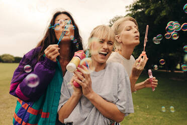 Friends hanging out together in a park, celebrating life and having fun. They are blowing bubbles, laughing, and socializing, all while wearing casual clothing and authentic smiles. This photo has intentional use of 35mm film grain. - JLPPF02286