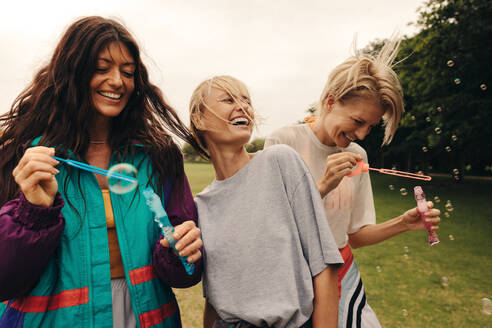 Three women enjoy a day in the park, celebrating friendship by blowing bubbles and playing games. Genuine laughter and smiles fill the air as they socialize and have fun together. This photo has intentional use of 35mm film grain. - JLPPF02288