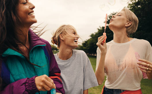 Girlfriends hang out in the park, enjoying a leisurely day. Friends playfully blow bubbles and laugh together, creating positive vibes and happy memories in their casual outfits. This photo has intentional use of 35mm film grain. - JLPPF02289