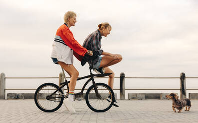 Female friends in their 30s have fun riding bikes along the seaside. They smile, laugh and enjoy together wearing casual clothes while socializing in a real and playful way. This photo has intentional use of 35mm film grain. - JLPPF02299