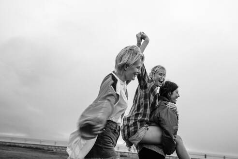 Three women having a blast together outdoors. They're in casual clothing, piggybacking and laughing with genuine joy. It's a real moment of friendship, celebration, and fun. This photo has intentional use of 35mm film grain. - JLPPF02300