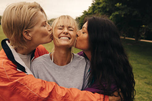 Two women embrace and kiss their friend on the cheeks while hanging out in a park. Group of middle aged friends having fun with each other outdoors. This photo has intentional use of 35mm film grain. - JLPPF02305