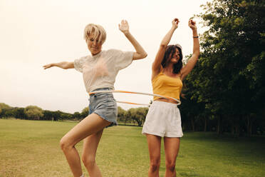 Two women enjoy a carefree summer day together, playing and dancing around in a hula hoop. Mature female friends, dressed in casual clothing, have fun while hanging out in a park. This photo features intentional film grain for artistic effect. - JLPPF02310