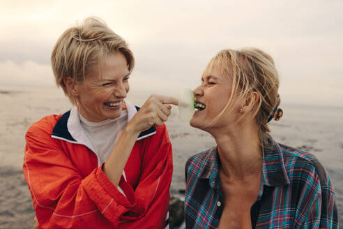 Playful woman popping her best friend's bubble gum, enjoying a moment of laughter and friendship vibes in the ocean breeze. Two best friends having fun together outdoors. This photo has intentional use of 35mm film grain. - JLPPF02324