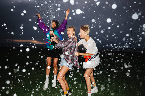 Three girlfriends cheerfully celebrate with champagne in the park, popping bubbles and screaming with laughter. Group of women having fun and enjoying their summer holiday together. This photo has intentional use of 35mm film grain. - JLPPF02325