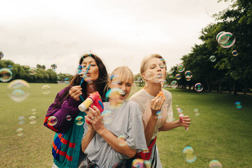 Group of women, best friends, enjoy a playful summer day in the park. Mature girlfriends blow vibrant bubbles and celebrate their long lasting friendship with smiles all around. This photo features intentional film grain for artistic effect. - JLPPF02327