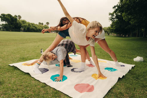 Female friends have a blast together in the park, playing a fun game of balance on a colourful mat. Group of women spending time together outdoors, enjoying authentic friendship vibes. This photo has intentional use of 35mm film grain. - JLPPF02328
