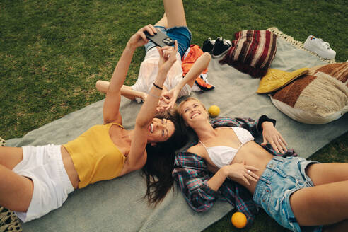 Kaukasische Freundinnen, die sich bei einem Obstpicknick amüsieren, Erinnerungen festhalten und Selfies machen, während sie auf dem Rasen des Parks liegen - JLPPF02332