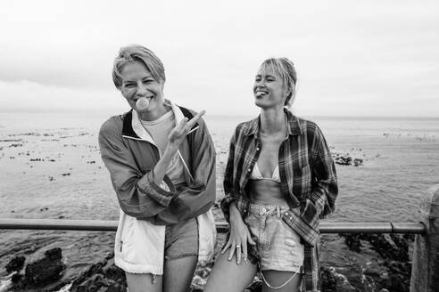 Cheerful female friends spend a carefree day by the sea promenade. They are happy and having fun, blowing bubbles with gum, enjoying the outdoors, and creating joyful moments together. This photo has intentional use of 35mm film grain. - JLPPF02337