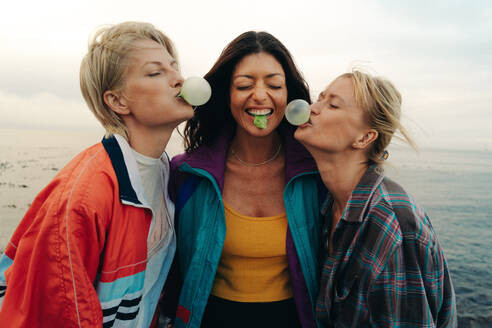 Group of female friends blowing bubbles with gum while hanging out together by the ocean. Three girlfriends laughing and having fun while surrounded by seaside breeze. This photo has intentional use of 35mm film grain. - JLPPF02339