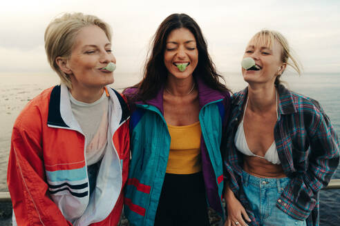 Group of female best friends having a fun day together by the ocean. Three happy women blowing bubbles and creating a playful atmosphere while enjoying the seaside breeze. This photo has intentional use of 35mm film grain. - JLPPF02340