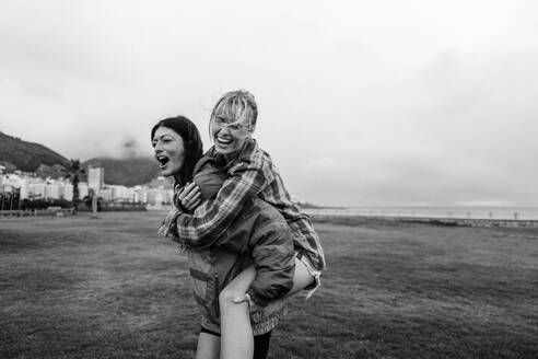 Two female best friends have fun together as they hang out in a park. Caucasian women piggybacking each other, enjoying authentic friendship vibes and creating memorable moments together. This photo has intentional use of 35mm film grain. - JLPPF02344