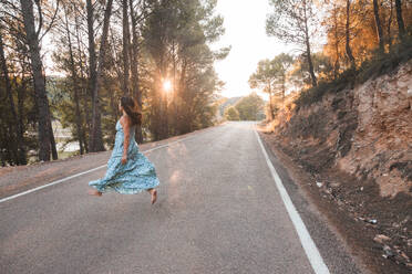 Side view of barefoot female traveler in blue summer dress jumping along empty asphalt road among trees during sunset time in Zaragoza city - ADSF44448