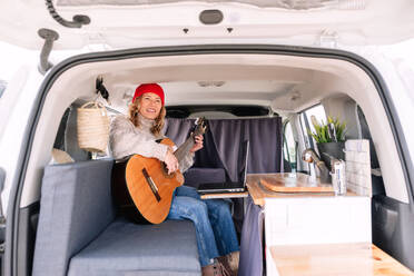 Cheerful young woman in casual clothes sitting at table with laptop and playing guitar in van - ADSF44455