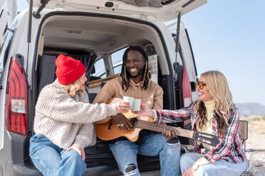 Cheerful friends sitting in van and playing guitar while spending time together on sunny day - ADSF44462