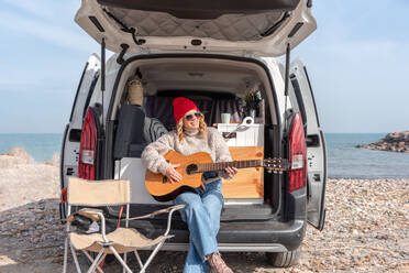 Full body of young smiling woman in casual clothes sitting in van and playing guitar while resting on beach near sea - ADSF44464