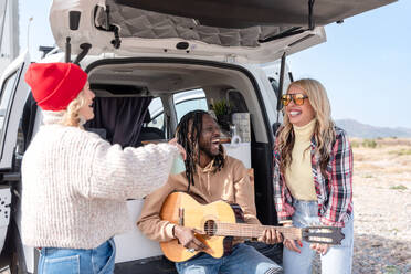 Cheerful friends sitting in van and playing guitar while spending time together on sunny day - ADSF44465