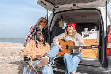 Cheerful friends sitting in van and playing guitar while spending time together on sunny day - ADSF44466