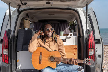 Happy black male with dreadlocks and sunglasses sitting in car with opened van door while playing guitar looking away - ADSF44467