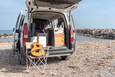 Side view of travel car parked on shore near sea with open trunk door while guitar left on chair on rocky beach in summer daylight - ADSF44469