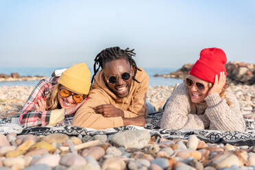 Smiling multiracial cheerful friends in sunglasses lying on blanket while relaxing on stony coast near sea in daylight - ADSF44470