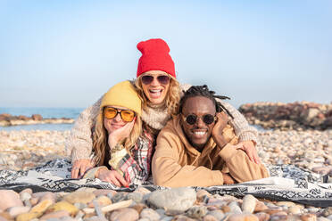 Smiling multiracial friends in sunglasses with red cap female on back lying on blanket and looking at camera while relaxing on stony coast near sea in daylight - ADSF44473