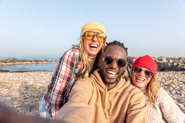 Group of cheerful young multiracial friends in sunglasses relaxing in summer daylight on pebbles beach near sea and looking away under blue sky - ADSF44474