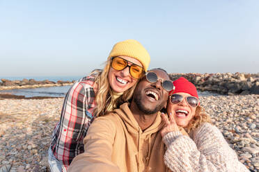 Group of cheerful young multiracial friends in sunglasses relaxing in summer daylight on pebbles beach near sea and looking away under blue sky - ADSF44475