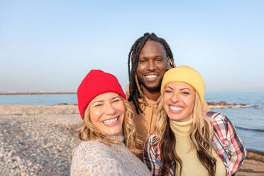 Positive young black male with cards hugging multiracial female friends in caps while smiling and looking at camera on sandy beach against sea and blue sky - ADSF44478