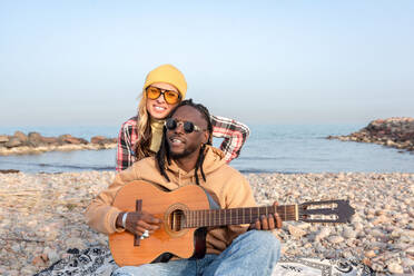 Happy young female embracing from behind cheerful black male looking away in sunglasses and playing acoustic guitar while relaxing on stony coast near sea in daylight - ADSF44481