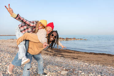 Cheerful young black male carrying piggyback both female friends showing victory sign and smiling while looking at camera and standing on stony coast near waving sea - ADSF44482