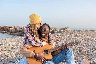 Happy young female embracing from behind cheerful black male looking away in sunglasses and playing acoustic guitar while relaxing on stony coast near sea in daylight - ADSF44484