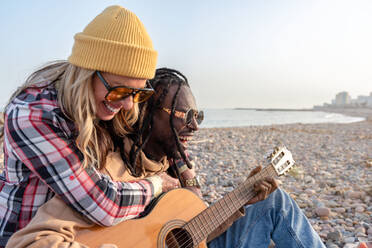 Happy young female embracing from behind cheerful black male looking away in sunglasses and playing acoustic guitar while relaxing on stony coast near sea in daylight - ADSF44485