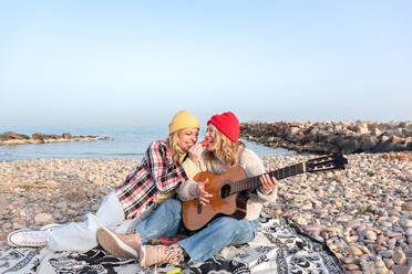 Happy young female in warm clothes hugging smiling friend looking at each other sitting on stony coast playing guitar near waving sea in daylight - ADSF44486