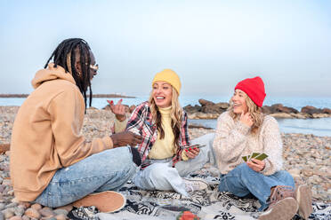 Side view of young females in caps laughing at black male with hand on forehead while sitting on blanket and playing cards in daylight against blue sky - ADSF44487