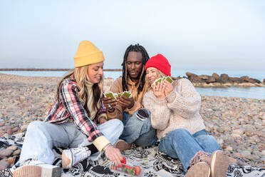 Upset black male showing cards to smiling diverse females in caps while sitting together on blanket with strawberries near sea water in daylight against cloudless blue sky - ADSF44489