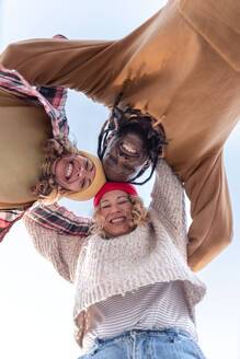 From below of cheerful group of multiracial friends with hands on shoulders smiling and looking down at camera against cloudless blue sky in summertime - ADSF44492